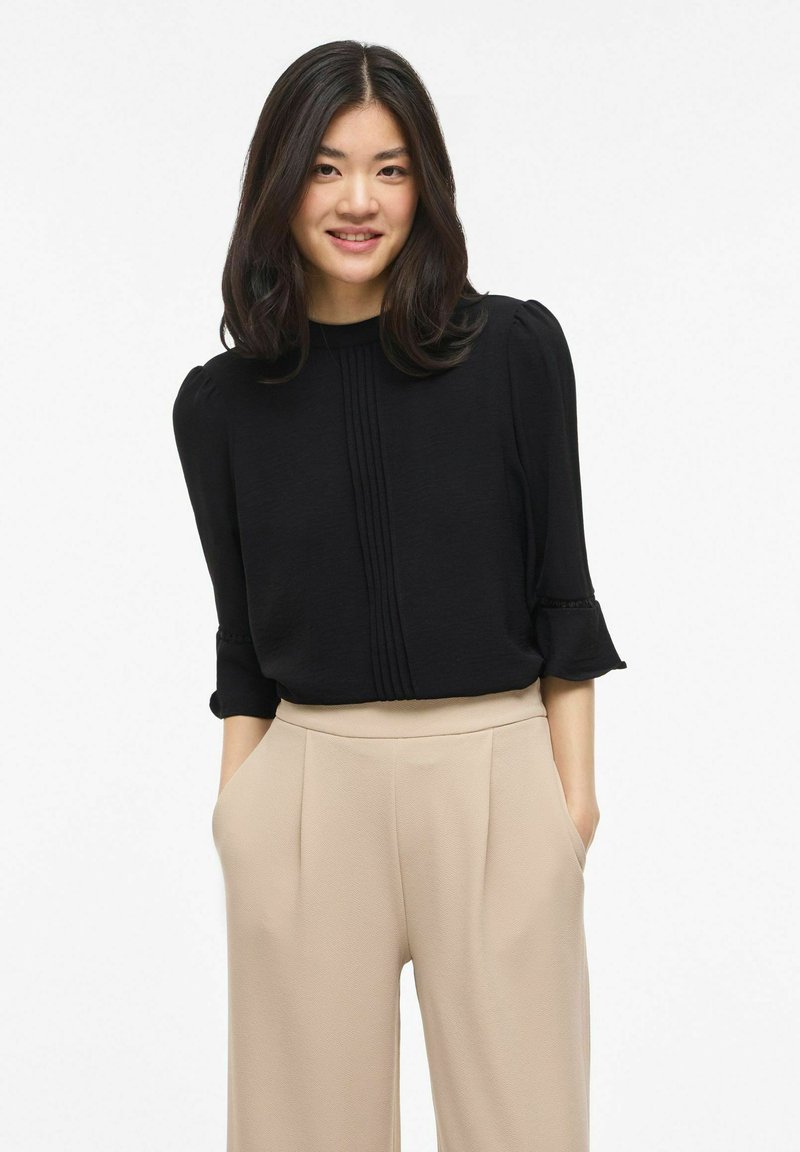 Jeune femme aux cheveux bruns mi-longs, portant un chemisier noir et un pantalon beige à jambes larges, mains dans les poches, souriant à la caméra.