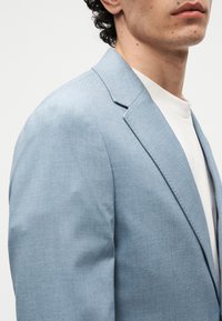 Κοντινό πλάνο ενός ατόμου που φοράει ανοιχτό μπλε σακάκι πάνω από λευκό μπλουζάκι με στρογγυλή λαιμόκοψη σε απλό φόντο.