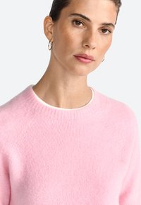 Vaaleanpunainen villapaita, joka on valmistettu pehmeästä, tekstuurilla varustetusta materiaalista. Ribbaettu kaula-aukko ja raglan-hihat. Minimalistinen muotoilu ilman kuvioita tai koristeita.