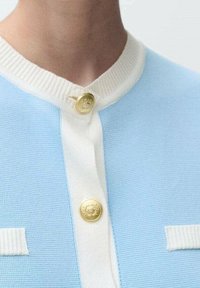 Cárdigan de punto azul claro con un cuello blanco acanalado, cierre con botones dorados y detalles blancos en los bolsillos. Textura suave en todo el prenda.