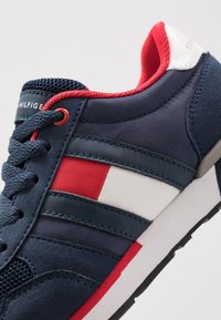 Mörkblå sneakers med mocka- och mesh-material, röda och vita accenter, texturerade överdrag och kontrasterande vit sula.