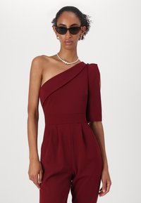 Combinaison bordeaux à une épaule avec un corsage ajusté, une manche structurée et un drapé subtil. Tissu lisse avec une taille cintrée et des jambes larges.