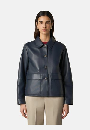 Donna che indossa una giacca di pelle blu navy con quattro bottoni e due tasche davanti, abbinata a pantaloni beige, in piedi davanti a uno sfondo semplice.