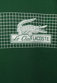 Tissu en maille verte avec un motif quadrillé blanc. Logo blanc central avec une illustration de crocodile et le texte « Le Club Lacoste » en dessous.