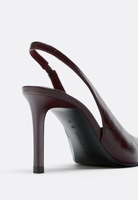 Scarpa a tacco alto slingback in pelle bordeaux con finitura lucida, punta affusolata e tacco sottile. Presenta un dettaglio di cinturino incrociato.