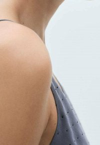 Zbliżenie na ramię i górną część pleców osoby w niebieskim, bezrękawniku z małymi czarnymi kropkami, na jednolitym jasnym tle.