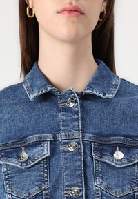 Blå denimjakke med krage, metallsnapper og to brystlommer. Stoffet viser et teksturert mønster og svak falming.