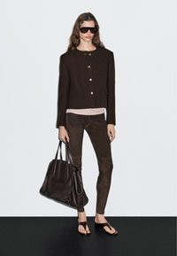 Vrouw in een donkerbruine knopenjasje, strakke broek, zonnebril, met een grote donkere leren schoudertas en open hakken.