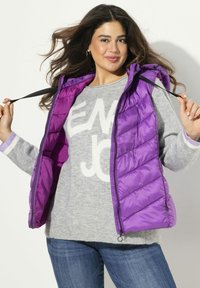 Gilet imbottito viola con cappuccio, caratterizzato da un design trapuntato diagonale. Indossato sopra un maglione grigio con un motivo di testo bianco. Jeans in denim visibili.