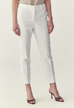 Kobieta w białych, dopasowanych spodniach do kostek z przednią plisą oraz brązowych, otwartych sandałach na obcasie, stojąca na jednolitym tle.