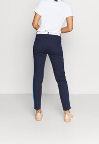 Marinblå slim-fitbyxor med en kontrasterande ljusblå sidremsa, med en slät yta och standard bakfickor, i kombination med beige skor.