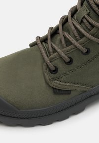 Botte en toile vert olive avec une semelle en caoutchouc, arborant des lacets marron, des œillets noirs et une petite étiquette de marque sur le côté.