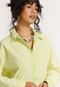 Lichtgroene overhemd met kraag, lange mouwen en subtiele textuur; gecombineerd met een meerkleurige kralenketting.