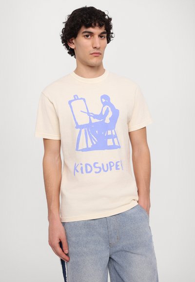 Ecrufarbenes Baumwoll-T-Shirt mit einem blauen Grafikdruck eines sitzenden Figuren, die malt, und dem Text "KIDSUPER" darunter. Kurze Ärmel, Rundhalsausschnitt.