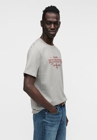 Γκρι βαμβακερό t-shirt με κοντά μανίκια, με κόκκινο τυπωμένο κείμενο. Φορεμένο με μπλε τζιν παντελόνι. Απλός, χαλαρός σχεδιασμός εφαρμογής.