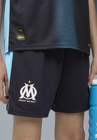 Juodi futbolo šortai su tekstūruotu paviršiumi ir mėlynomis akcentais, su baltu logotipu ir auksinėmis raidėmis. Įtrauktas auksinis Puma autentiškumo ženklelis.
