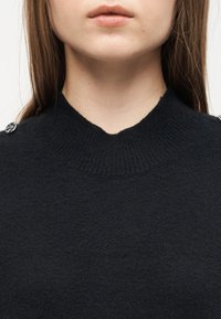 Gros plan sur une personne portant un pull à col roulé noir côtelé avec des détails de boutons sur les épaules.