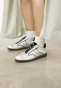 White sneakers with black accents, featuring a smooth leather upper, contrasting stitching, and a patterned rubber sole. White ribbed socks visible.