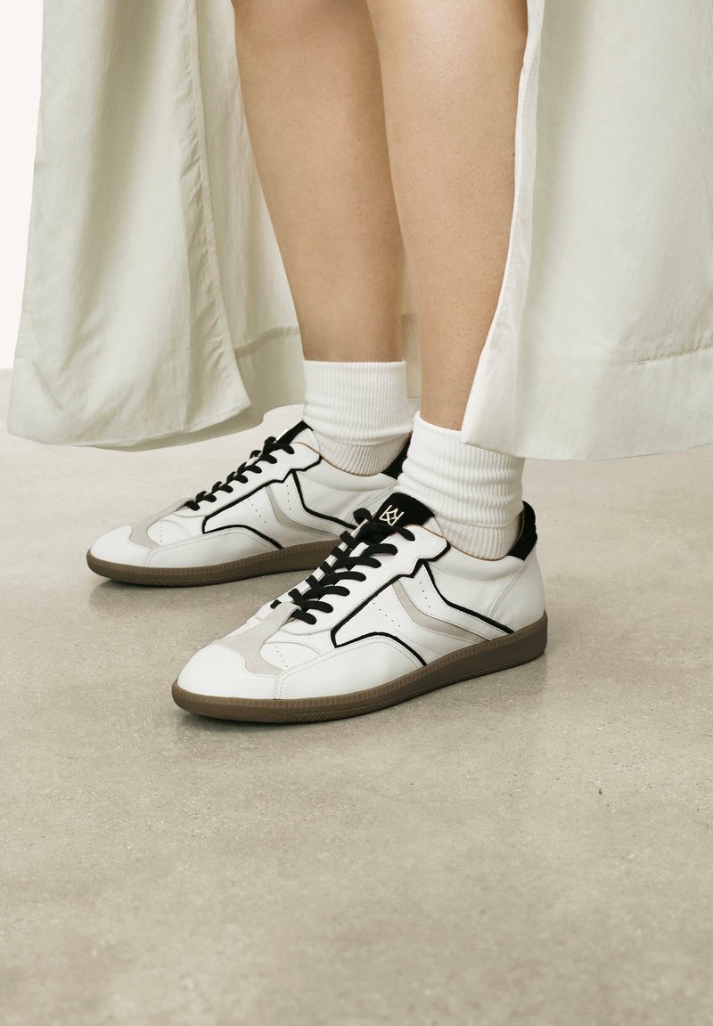White sneakers with black accents, featuring a smooth leather upper, contrasting stitching, and a patterned rubber sole. White ribbed socks visible.