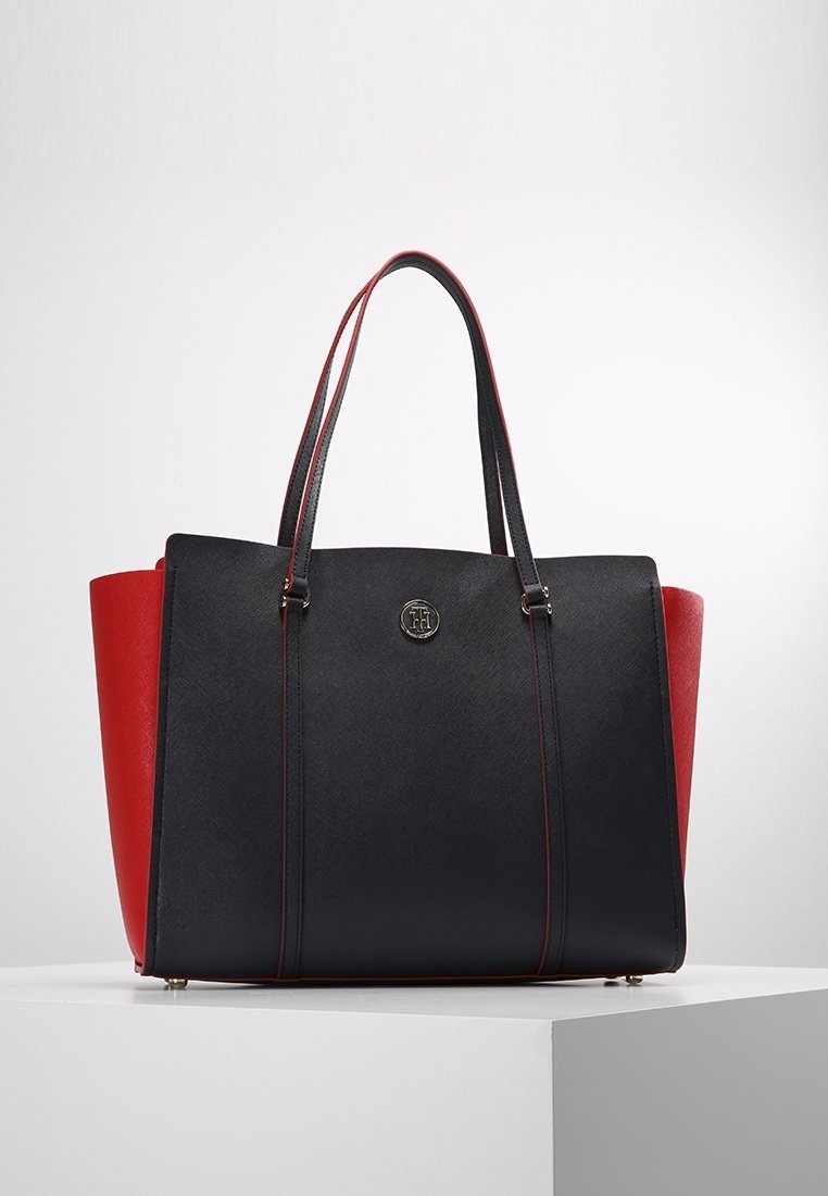 Sac à main noir et rouge avec deux poignées, panneaux latéraux triangulaires et un logo métallique rond au centre avant, posé sur une surface blanche.