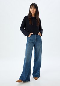 Marineblauer Strickpullover, weit geschnittene blaue Jeans und beige, spitz zulaufende Schuhe. Das Outfit zeichnet sich durch eine entspannte Passform und strukturierte Stoffe aus.