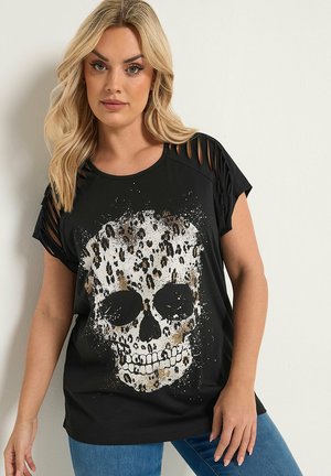 Donna bionda che indossa una maglietta nera con maniche sfilacciate e una grande grafica bianca e marrone con teschio a stampa leopardata, abbinata a jeans blu.