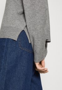 Pull en tricot gris avec des poignets côtelés et des fentes sur les côtés, porté sur un jean en denim bleu foncé avec des coutures de poche visibles.