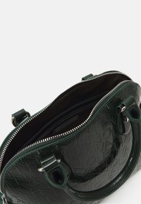 Glansig mörkgrön handväska med präglad logotyp, dubbla handtag upptill och dragkedjestängning som avslöjar ett brunt innervärde med fickor.