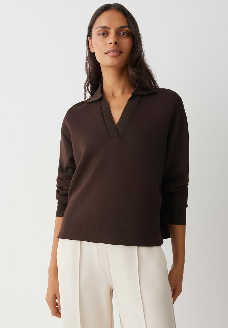 Brauner Pullover mit V-Ausschnitt und geripptem Kragen, gestaltet mit langen Ärmeln und legerer Passform, kombiniert mit hellfarbigen Hosen.