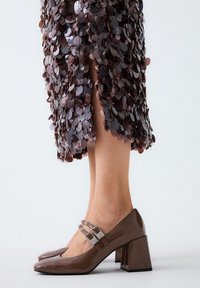 Zapatos de charol marrón con tacón cuadrado, puntera cuadrada y doble tira adornada con detalles de strass. Combinados con una falda con acentos de lentejuelas.