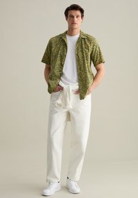 Camicia a maniche corte con motivo verde sopra una maglietta bianca a girocollo, abbinata a pantaloni bianchi a vestibilità rilassata e sneakers bianche.