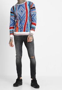 Pull coloré à col rond avec des motifs géométriques complexes en bleu, rouge et noir, associé à un jean skinny noir déchiré et des baskets.
