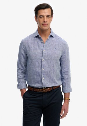 Mężczyzna o ciemnych włosach, ubrany w jasnoniebieską, zapinaną na guziki koszulę z długim rękawem, ciemne spodnie, brązowy pleciony pasek i beżowy zegarek na rękę.