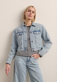 Lyseblå kort denimjakke med to brystlommer, sølvknapper og broderte detaljer. Brukt over en svart og hvit stripete topp.
