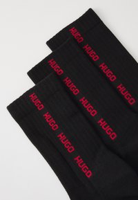 Trois paires de chaussettes noires côtelées avec le logo rouge "HUGO" répété près du haut du revers, posées à plat sur une surface blanche.