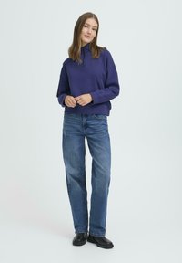 Vijoličen pulover z visokim ovratnikom, v kombinaciji s svetlo modrimi ravnimi hlačami in črnimi čevlji. Mehak material, sproščen kroj, minimalističen dizajn.