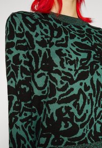 Pull noir et vert à motif, présentant un design en tricot texturé avec un motif animalier et un col côtelé.