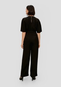 Zwarte jumpsuit met een ontspannen pasvorm, korte mouwen, elastische taille en een ritsdetail op de rug. Glad materiaal en een wijde pijpdesign.