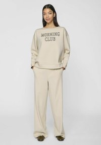 Sudadera beige con el texto "MORNING CLUB", combinada con pantalones de pierna ancha a juego. Tejido suave, corte relajado y mangas con puños.