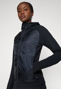 Veste zippée noire avec un panneau avant structuré et texturé. Comprend une capuche, des manches longues, et un design ajusté avec des poches latérales.