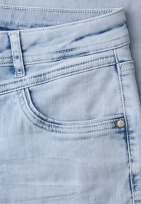 Gros plan sur un jean en denim bleu clair montrant la poche avant, la passants de ceinture, les détails des coutures et un petit rivet métallique sur le bord de la poche.