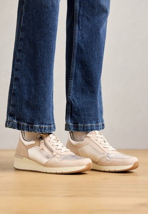 Baskets beige clair avec une finition texturée, dotées d'une fermeture éclair sur le côté, de lacets blancs et d'une semelle marron contrastante. Portées avec un jean bleu à jambes larges.