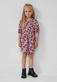 Junges Mädchen mit langen blonden Haaren, das ein pinkes Kleid mit Blumenmuster und schwarze Schnürstiefeletten trägt, steht gegen eine schlichte helle Wand.
