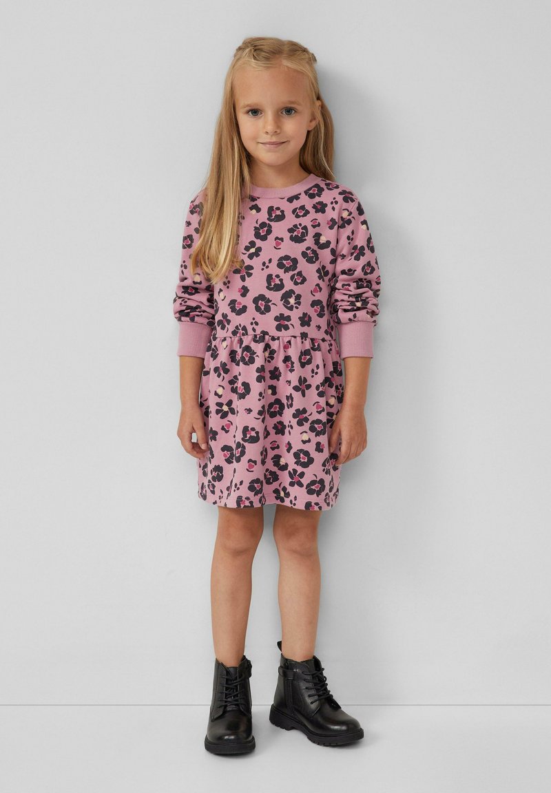 Junges Mädchen mit langen blonden Haaren, das ein pinkes Kleid mit Blumenmuster und schwarze Schnürstiefeletten trägt, steht gegen eine schlichte helle Wand.