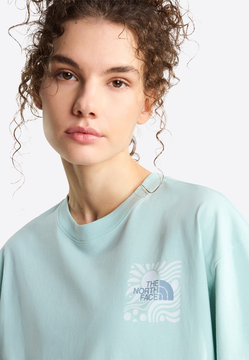 Junge Frau mit lockigem Haar, die einen hellblauen The North Face Pullover trägt und mit neutralem Gesichtsausdruck direkt in die Kamera schaut.