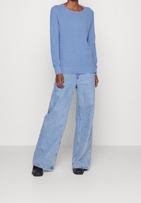 Pull texturé bleu clair avec manches raglan, associé à un jean en denim bleu clair à jambes larges. Des chaussures noires à bout carré complètent la tenue.