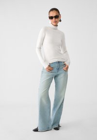 Bela turtleneck pulover v kombinaciji s svetlo modrimi širokimi hlačami. Dodatki vključujejo temne sončne očala in velike okrogle uhane. Črne čevlje.