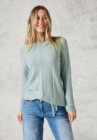 Haut long à manches longues vert clair texturé avec un ourlet droit, comportant une fente et des liens à la taille, associé à un jean en denim bleu.
