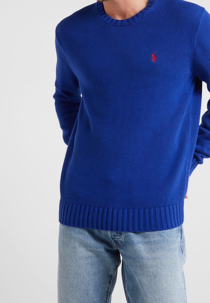Homme portant un pull bleu en maille avec un petit logo rouge sur la poitrine et un jean denim bleu clair sur un fond uni.