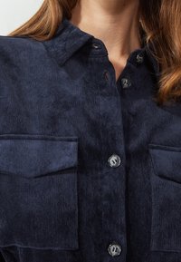 Navy corduroy shirt with a collared neck, two chest pockets, and black buttons. Texture appears soft and ribbed.